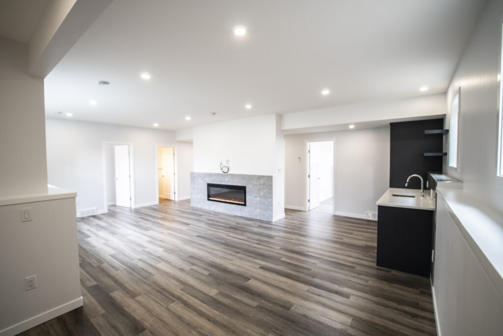 Modern, spacious living room with a kitchen to the right, featuring dark cabinetry and white countertops. Room includes a fireplace set in a white wall and wooden flooring throughout.