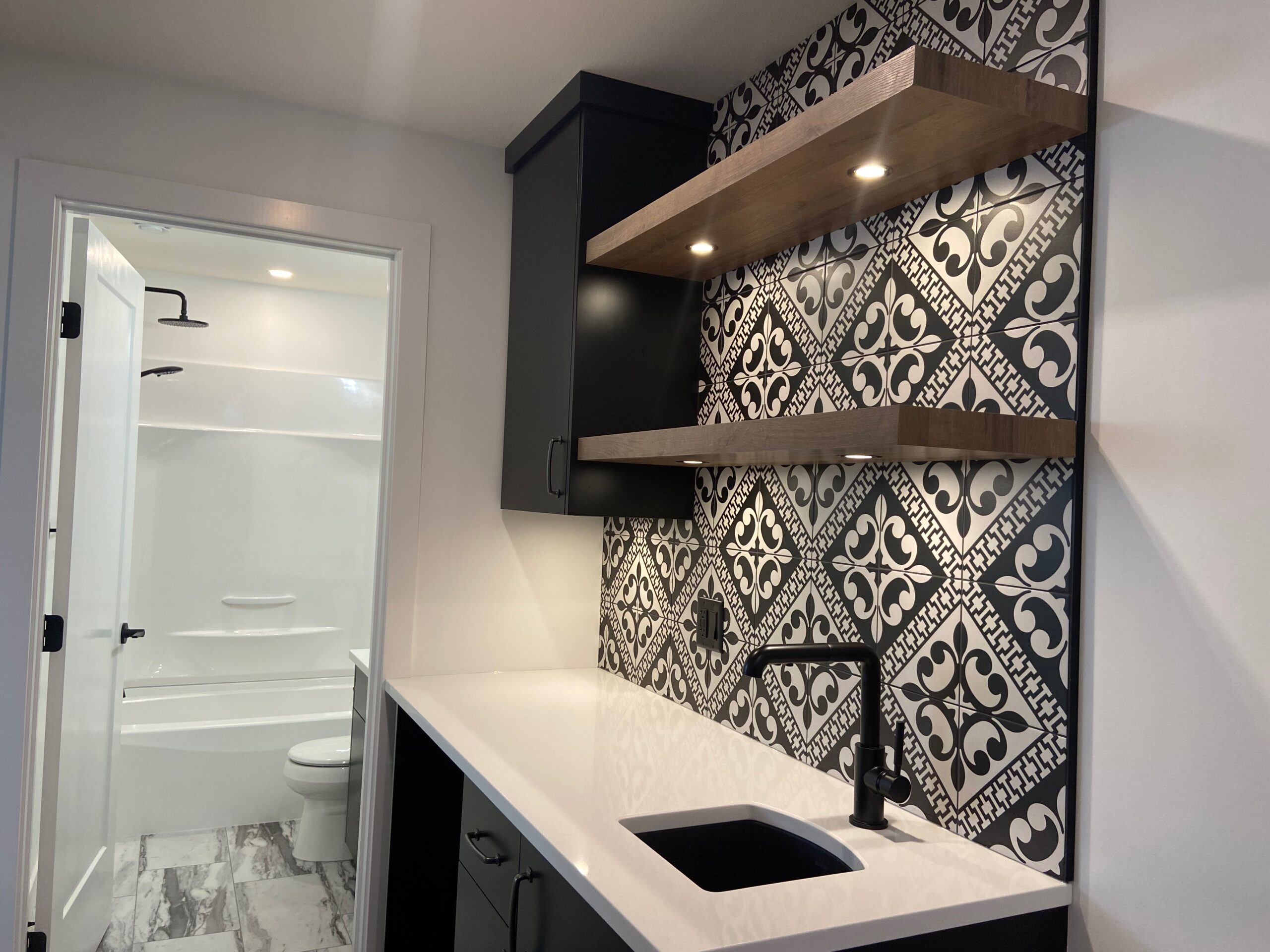 A modern bathroom features a black and white patterned tile backsplash, two floating wooden shelves, a white countertop with a black under-mount sink, black cabinetry, and a sleek black faucet. There is a glimpse of a white bathtub and toilet through an open door.