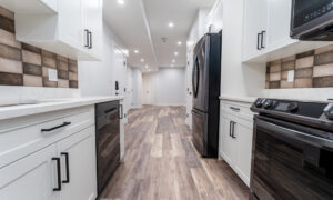 A modern kitchen interior featuring white cabinetry, striped backsplash tiles, and stainless-steel appliances. The floor is covered with wood-like tiles extending into a well-lit hallway.