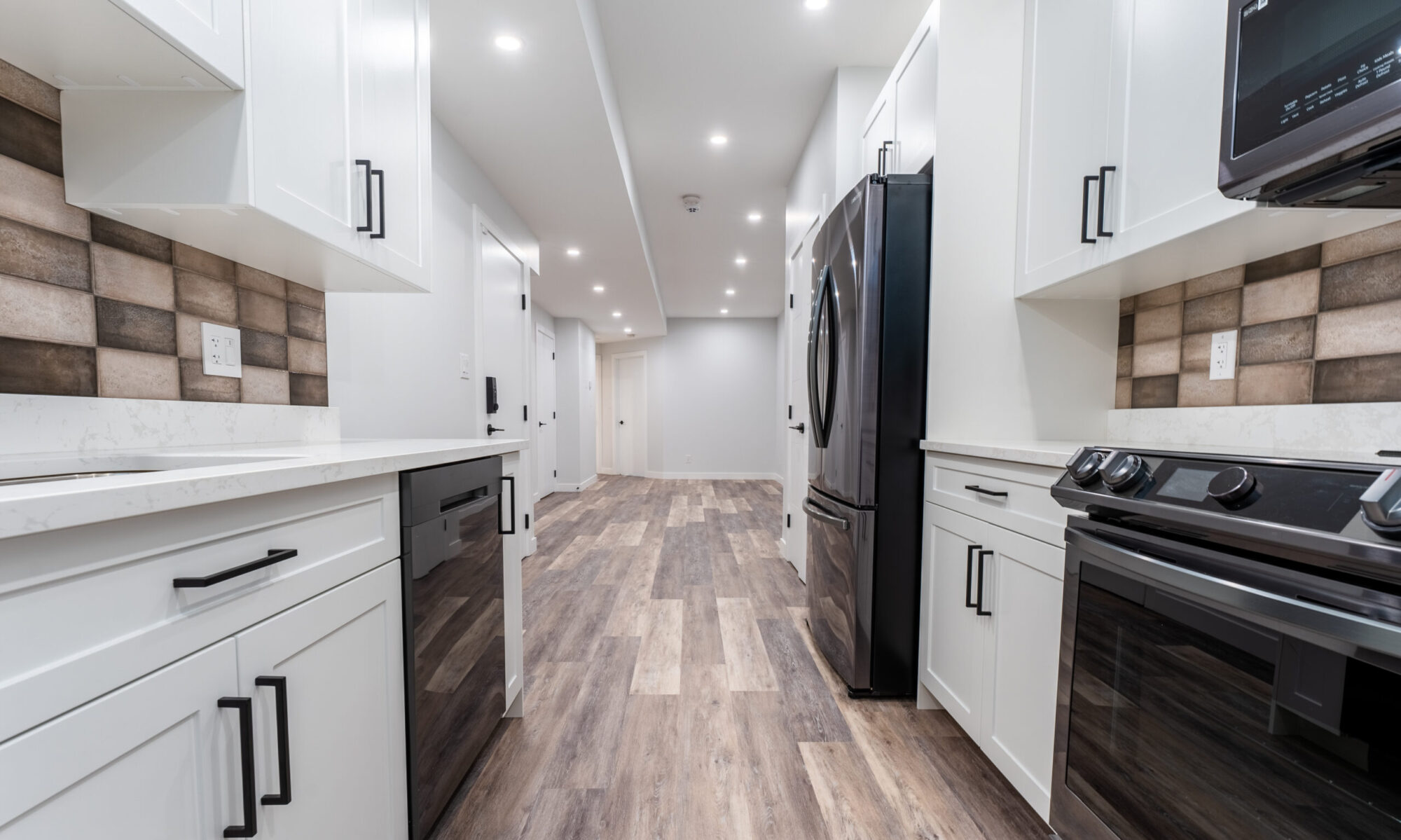 A modern kitchen interior featuring white cabinetry, striped backsplash tiles, and stainless-steel appliances. The floor is covered with wood-like tiles extending into a well-lit hallway.